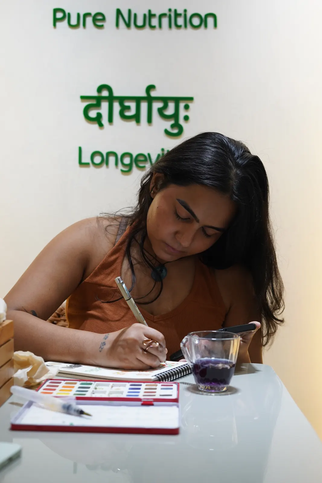 A guest sketching at a café table with a glass of herbal tea, under the Satvik Salaad brand wall